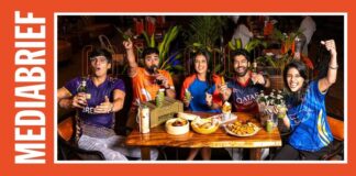 Five friends at a restaurant table raise drinks in a toast, with bowls of snacks and bottles in view, framed by an orange border and a vertical 'MEDIA BRIEF' label on the left.