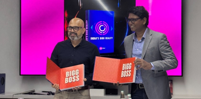 Two men on a stage hold open red 'Bigg Boss' booklets in front of a pink-screen backdrop.