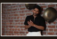 Guru Randhawa contributes Rs 5 lakh towards Assam flood relief efforts Portrait of a man in a black shirt and white pants posing with hands together in front of a brick wall and two round metal discs; vertical 'MEDIA BRIEF' text on the left.