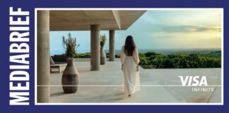 Woman in a white dress walks toward the ocean from a modern terrace; branding shows MEDIABRIEF on the left and VISA INFINITE on the right edge.