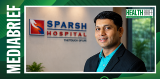 Professional portrait of a man in a blue shirt and dark suit standing in front of a Sparsh Hospital sign, with a Health Brief logo in the corner.